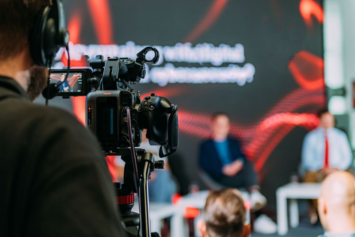 A video camera operator films a panel discussion on stage with a red digital backdrop at a professional event.