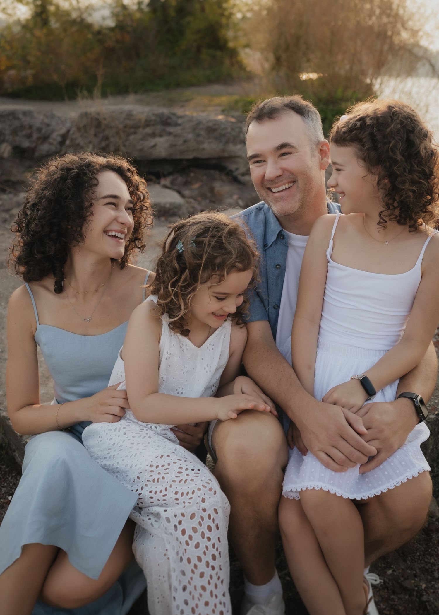 A happy family of four sits outdoors, smiling and laughing together in warm golden light near a rocky waterside.