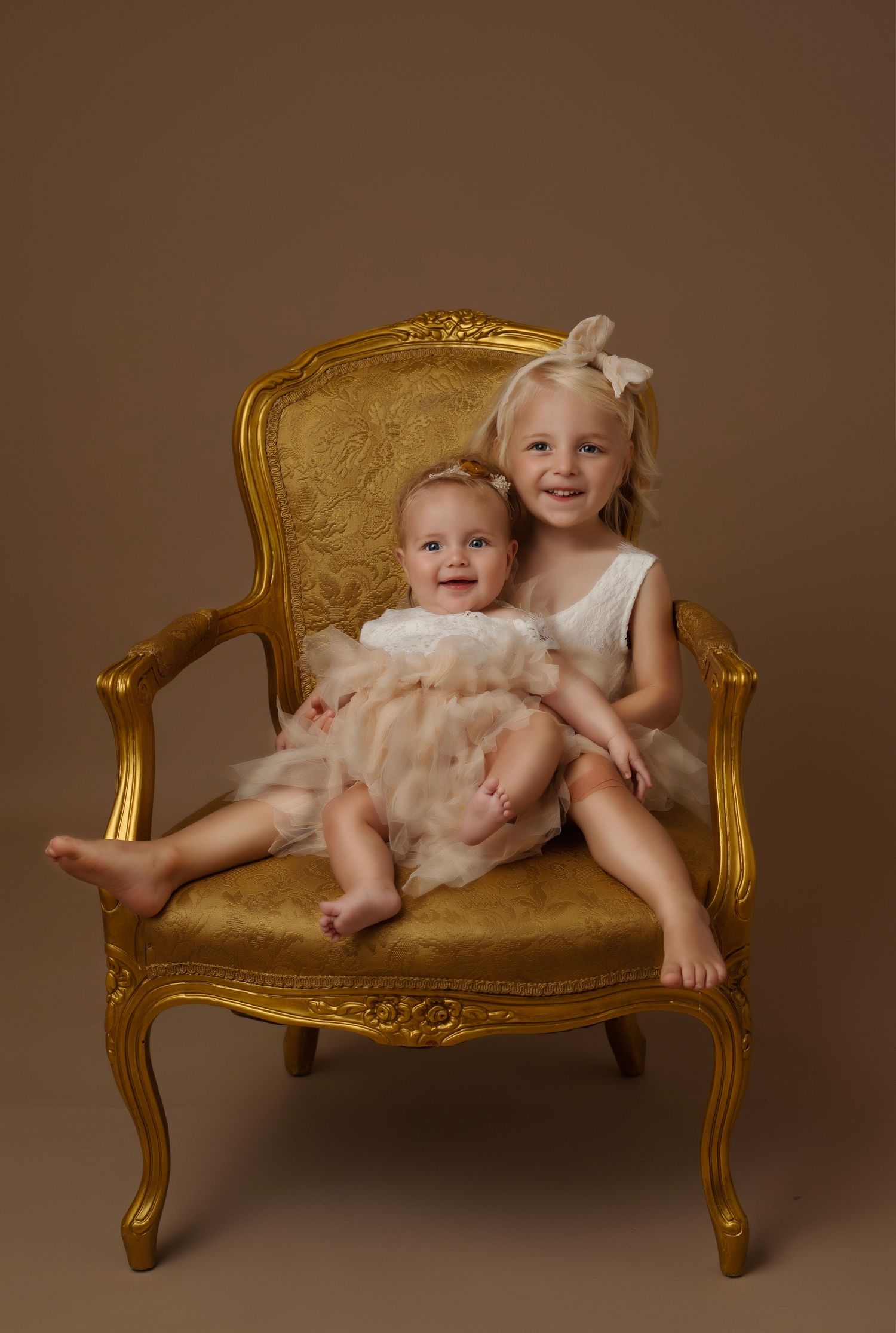 Two children posed on ornate gold vintage chair in cream colored outfits.