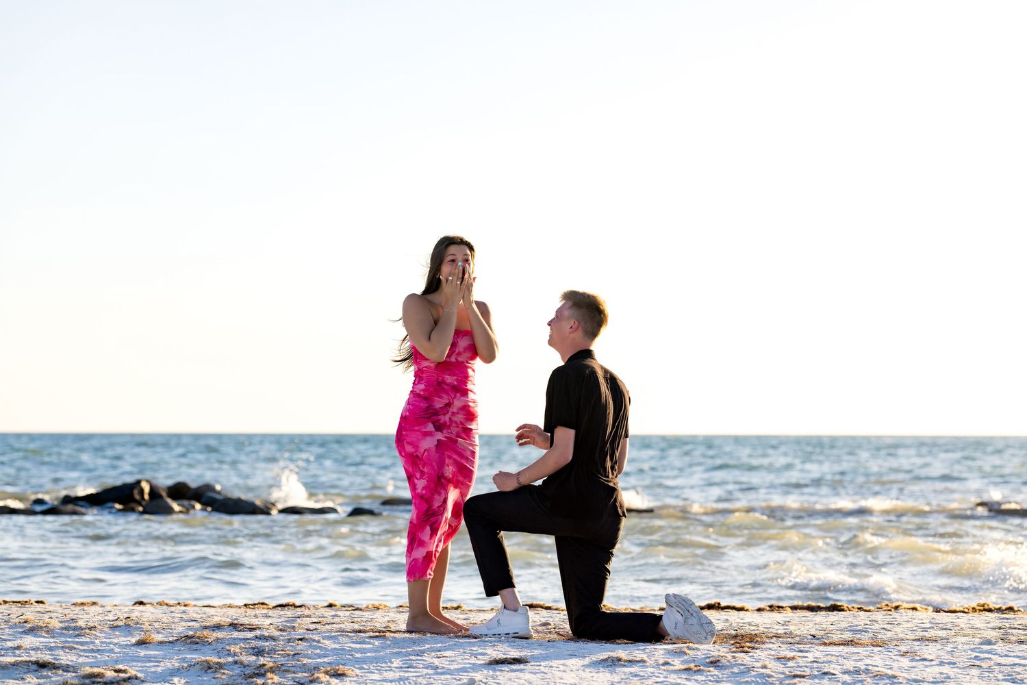 Man proposes to woman on a sandy beach at sunset, she reacts with surprise while he kneels holding a ring box.