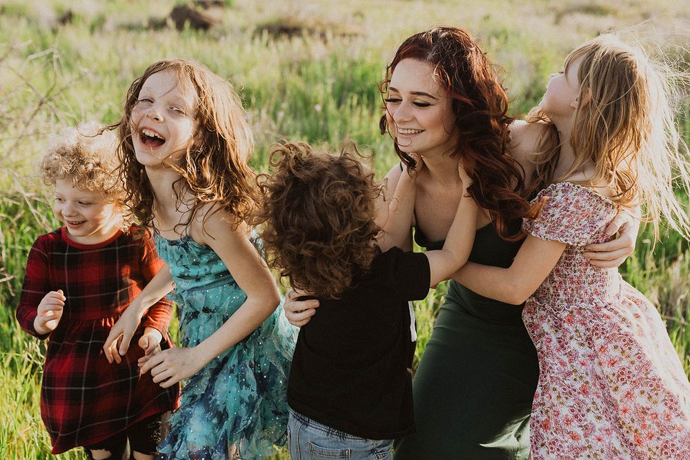 A joyful woman with red hair laughs with four young children in a sunny green field during a family photo session.