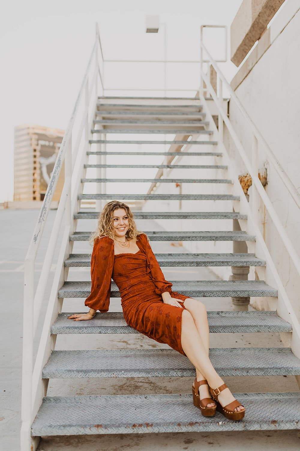 Woman in rust-colored midi dress and platform sandals sitting on metal stairs outdoors, smiling.