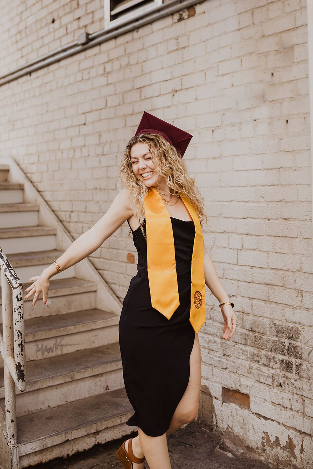 Smiling graduate in black dress with gold stole and maroon cap poses on outdoor stairs against brick wall.
