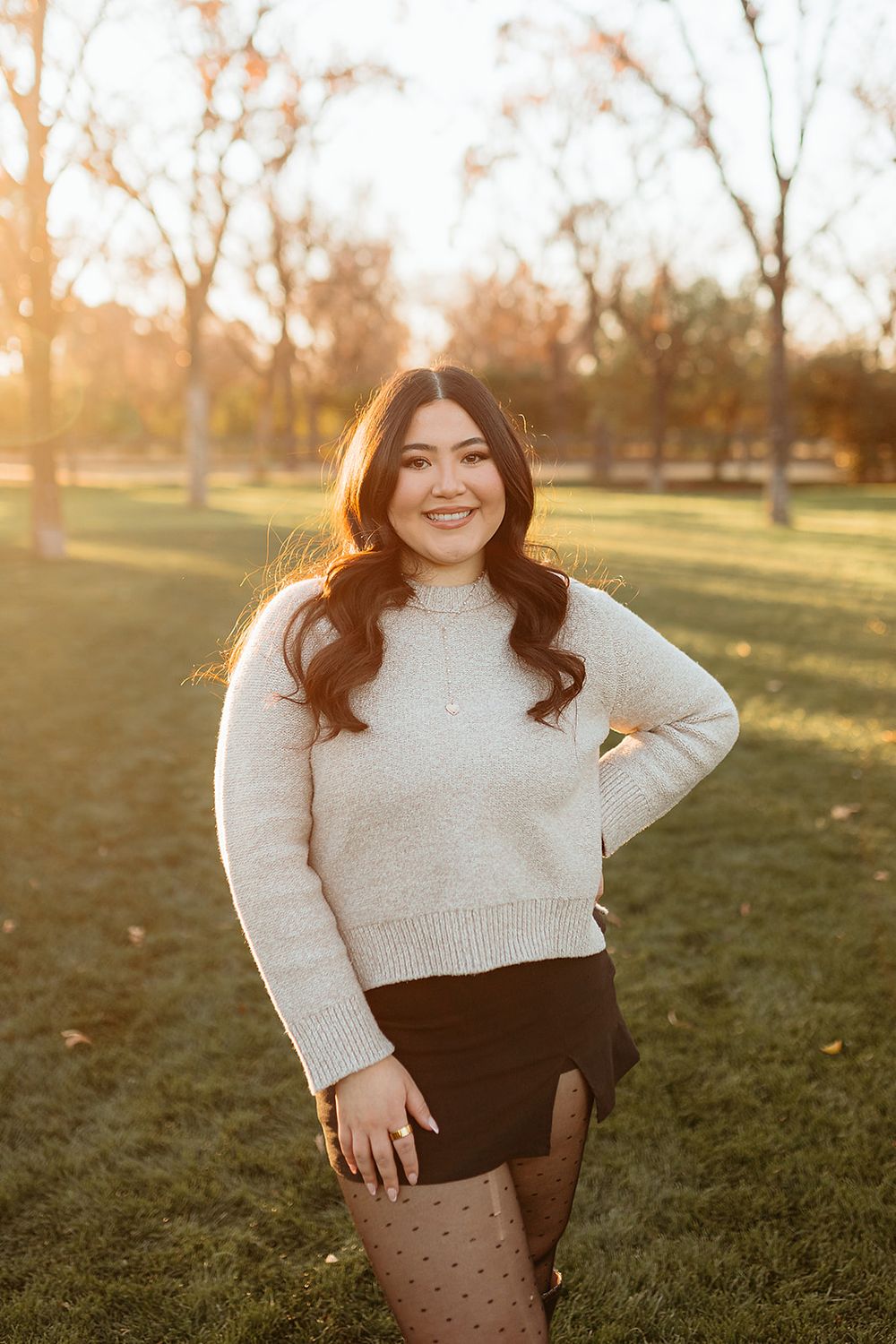 Young woman in cream sweater and black mini skirt posing in a sunny park during golden hour.