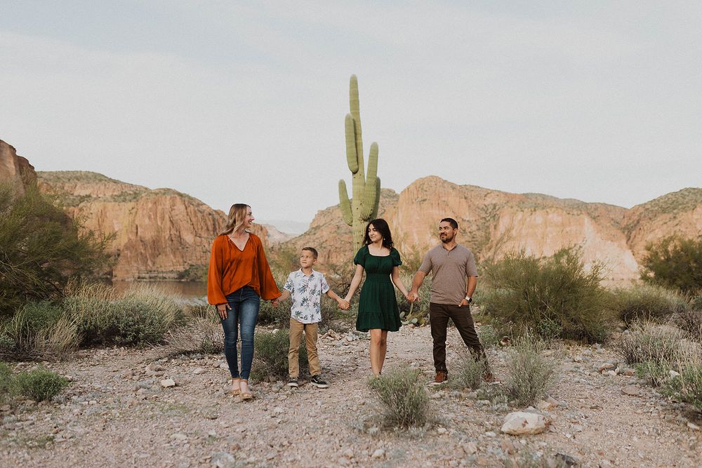 Family of four holding hands walking in Arizona desert with saguaro cactus and red rock mountains backdrop.