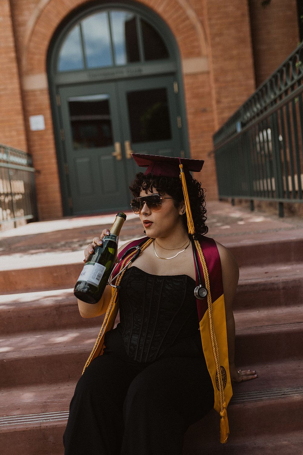 Graduate in cap and gold stole holds wine bottle on brick steps, wearing black corset and sunglasses.