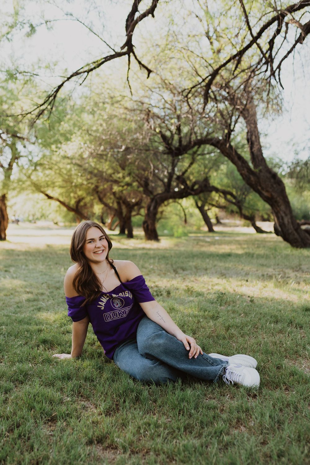 Young woman in purple off-shoulder top and jeans sitting on grass in a sunlit park with trees.