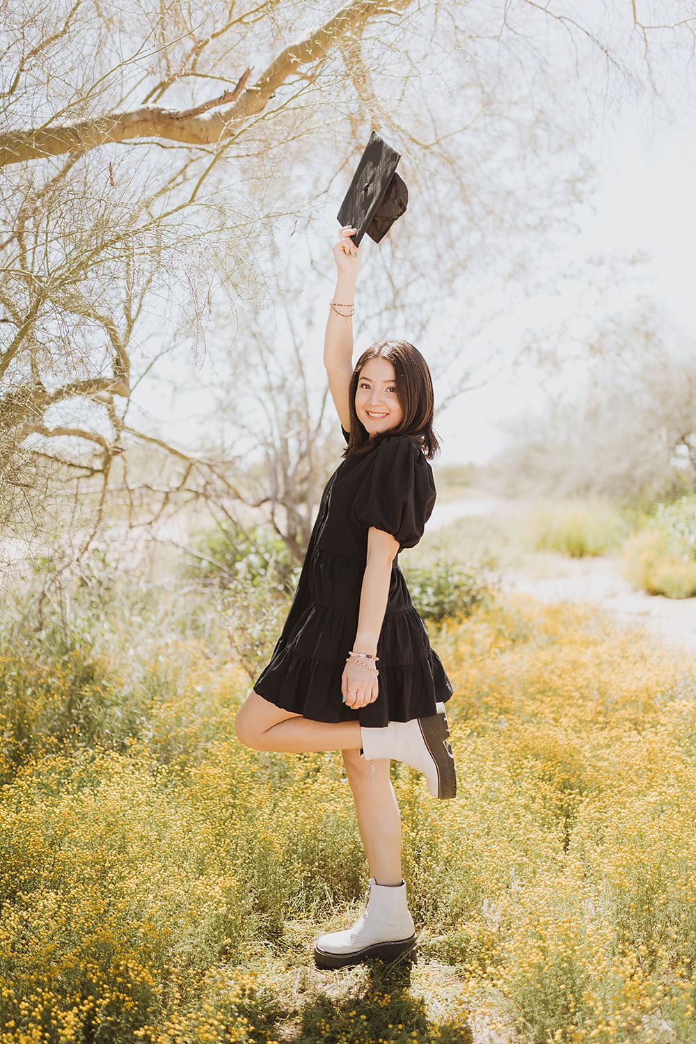 Young woman in black dress holding graduation cap aloft among yellow wildflowers, celebrating academic achievement outdoors.
