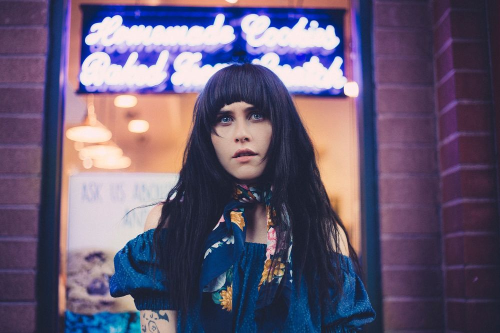 Woman with dark hair in blue floral jacket poses before neon sign reading Homemade Cookies at night.