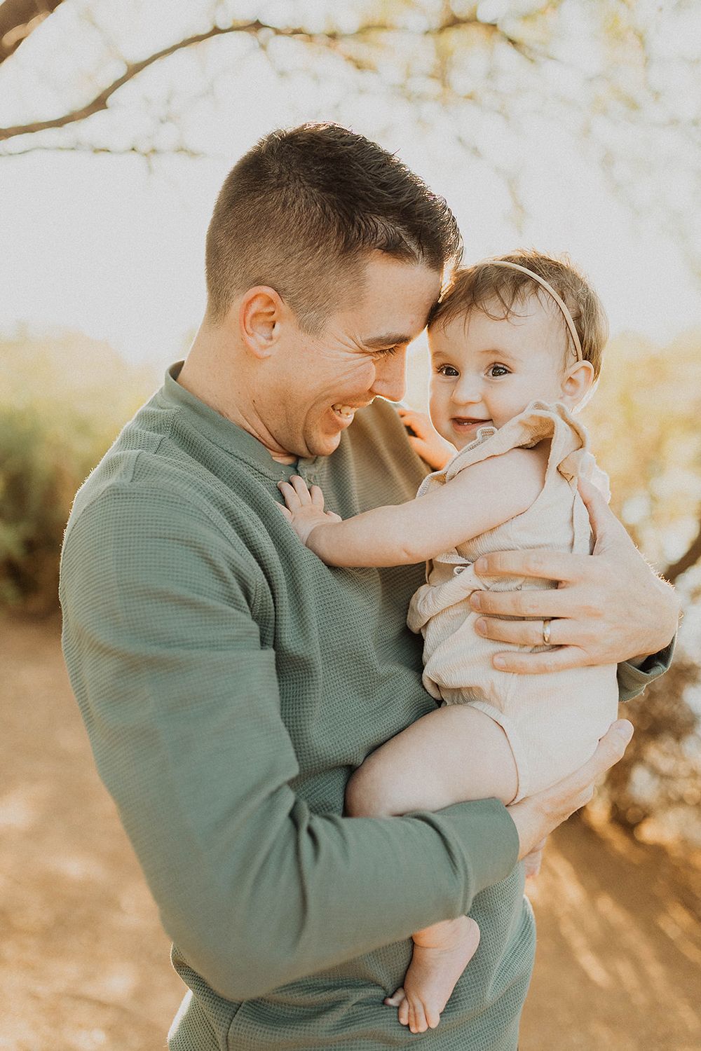Smiling man in green sweater holding a happy baby girl outdoors in warm golden sunlight.