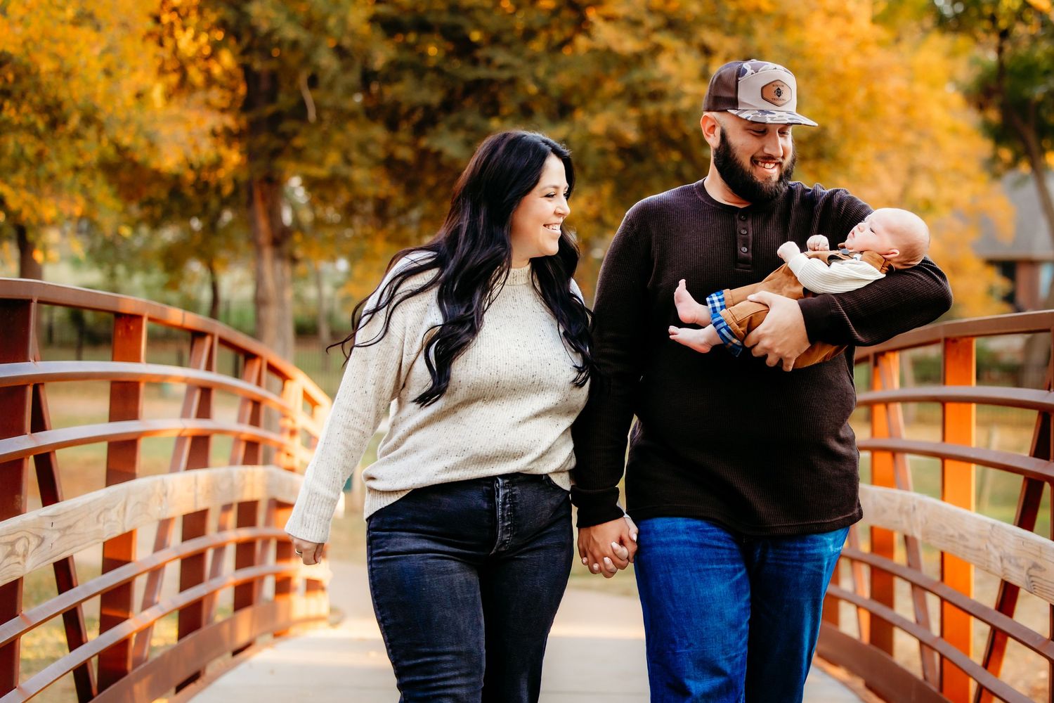 A couple walks hand-in-hand on a wooden bridge at Bear Creek Park in Keller, the man cradling a newborn baby amid golden autumn foliage.