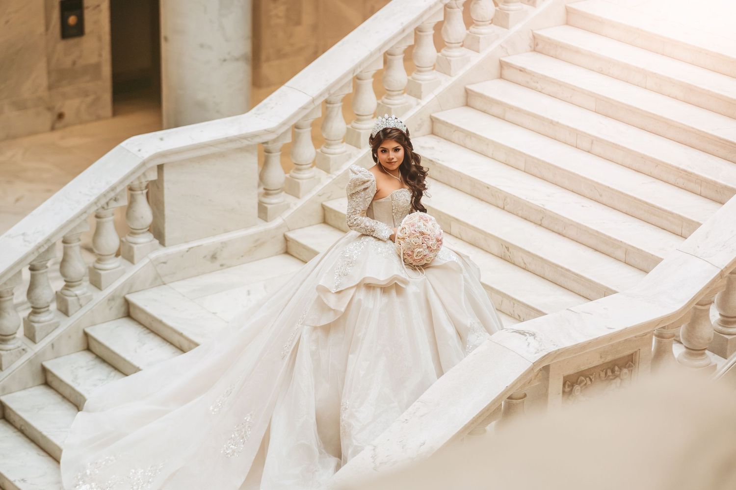 Bride in elegant white ball gown with long train poses on marble staircase wearing tiara and holding bouquet.