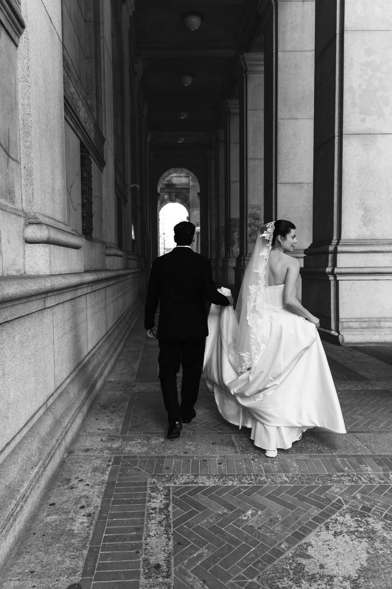 Black and white photo of bride and groom walking hand in hand through elegant stone archway corridor.