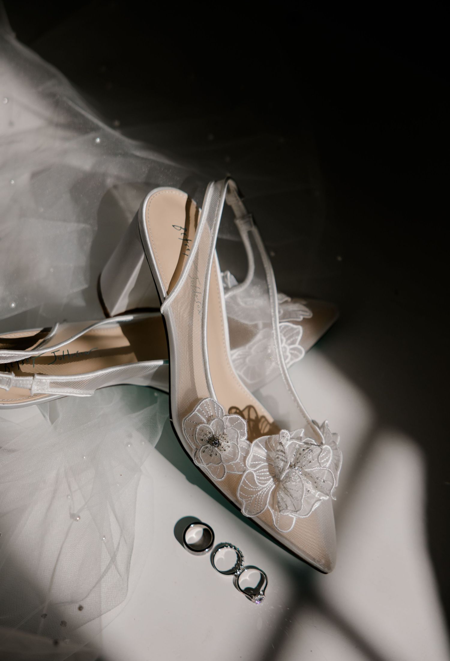 Elegant white lace bridal heels with floral detail beside wedding rings on a soft draped veil background.