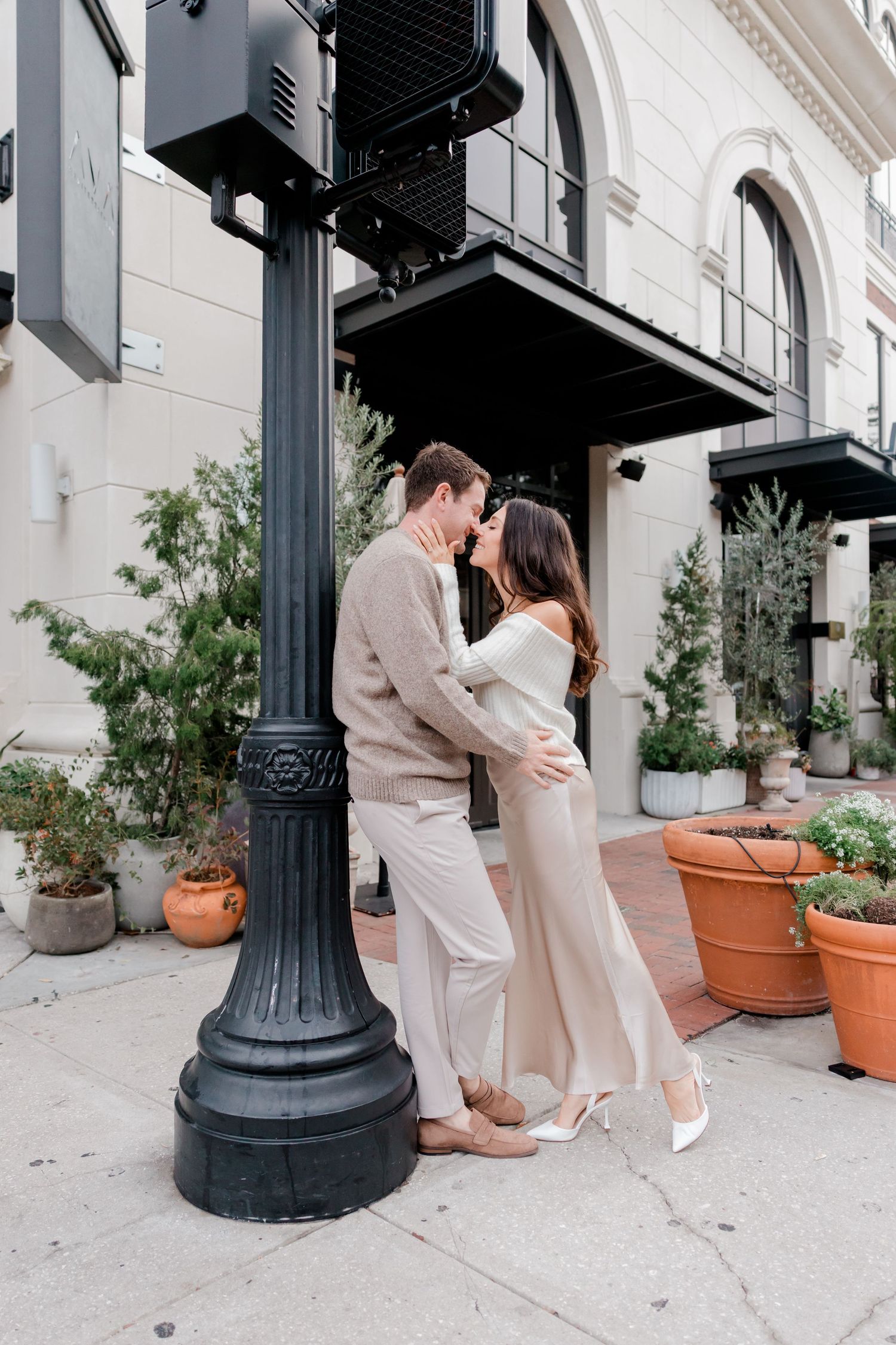 Couple sharing a romantic moment by a black street lamp outside a building with potted plants.