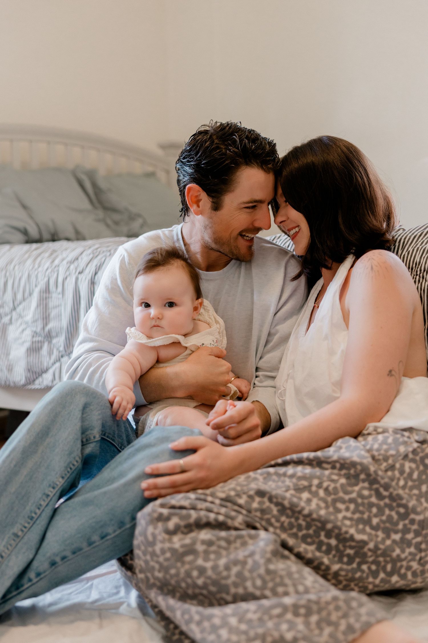Happy young couple sitting on bed smiling at each other while holding their adorable baby in a cozy bedroom.