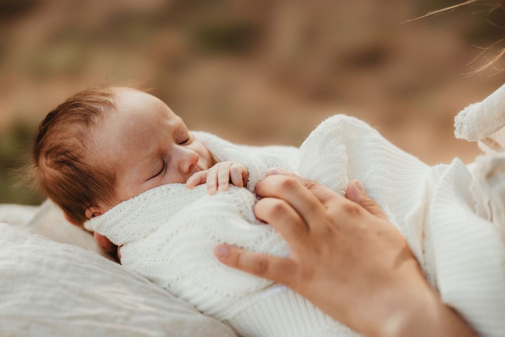 Peaceful newborn baby sleeping wrapped in white knit blanket, cradled gently in mother's arms outdoors.