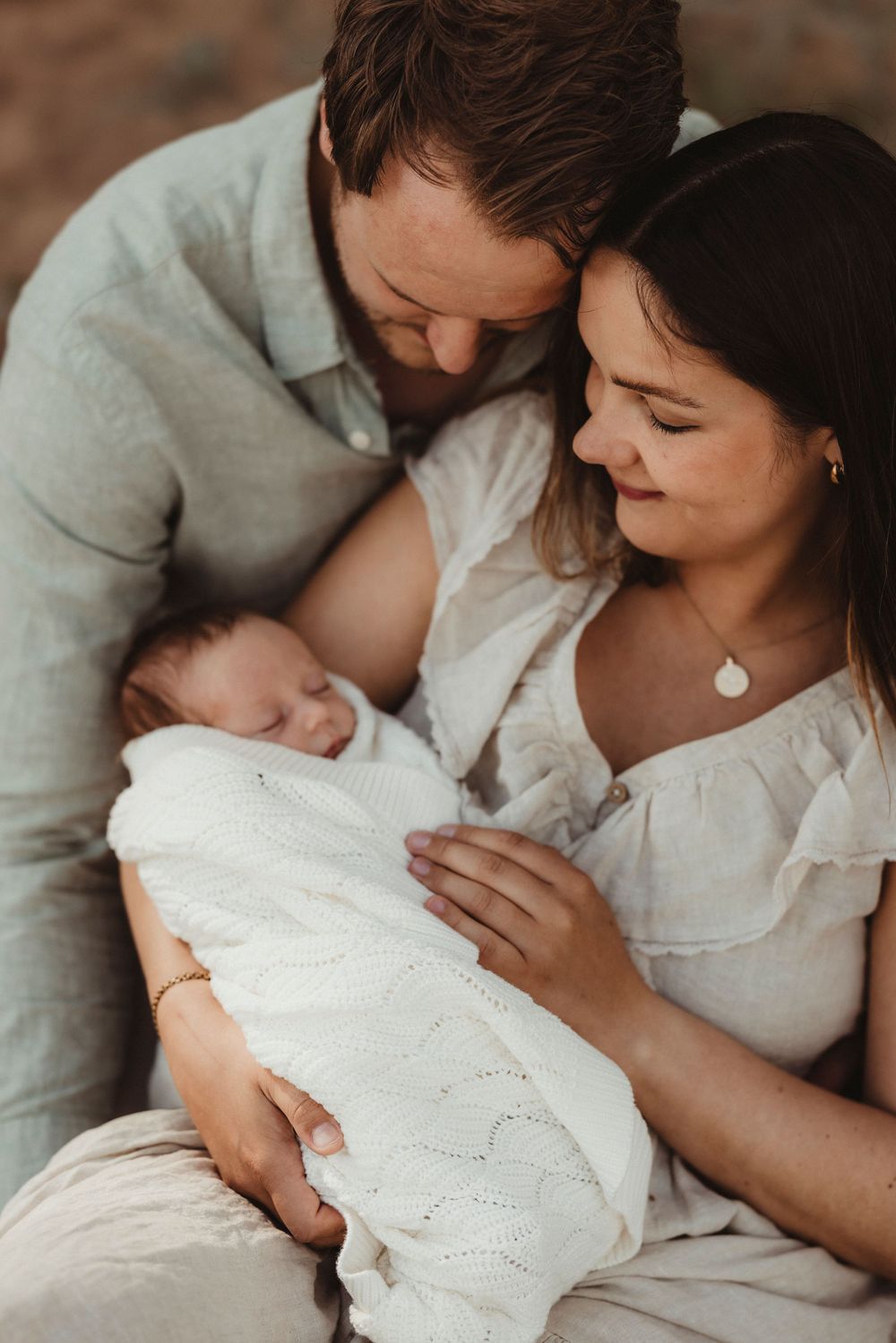 A couple tenderly gazes at their sleeping newborn wrapped in a white knit blanket during an outdoor family photo session.