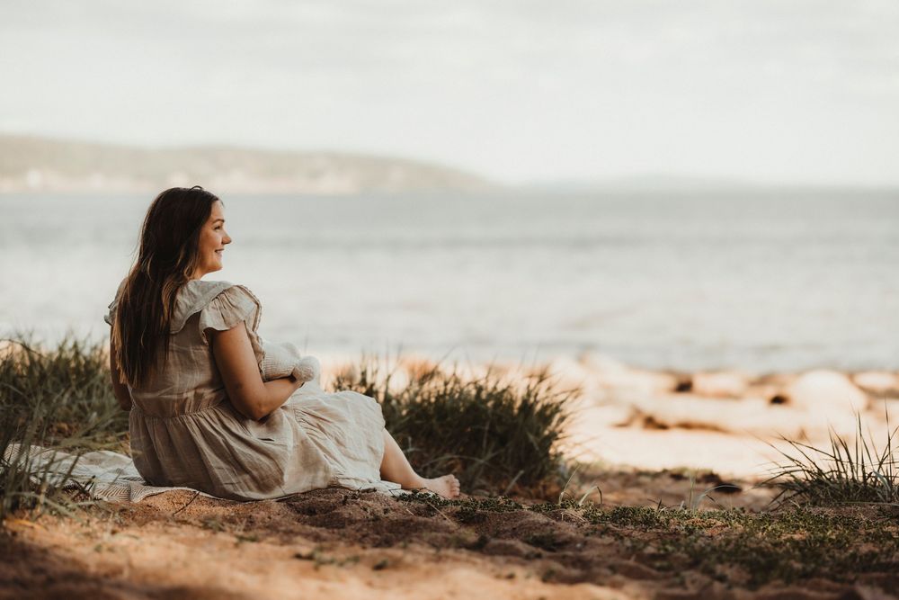 Woman in floral dress sitting on sandy shore gazing at ocean horizon during golden hour.