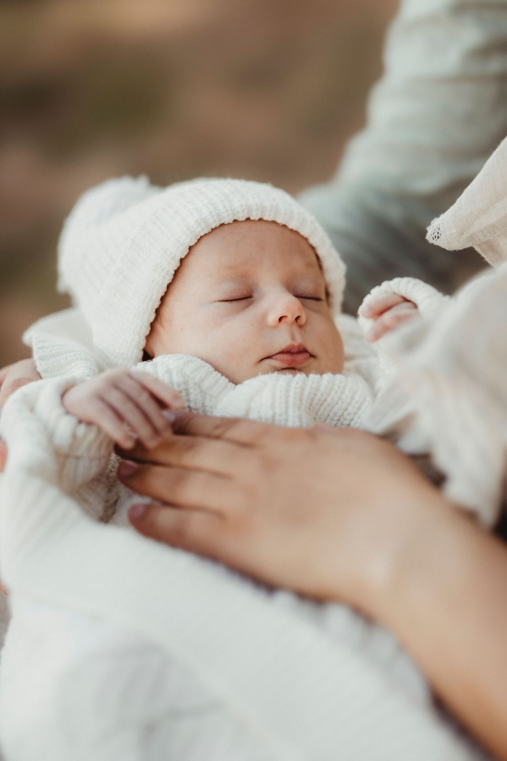 Newborn baby sleeping peacefully in white knit hat and outfit, cradled gently in parents arms outdoors.