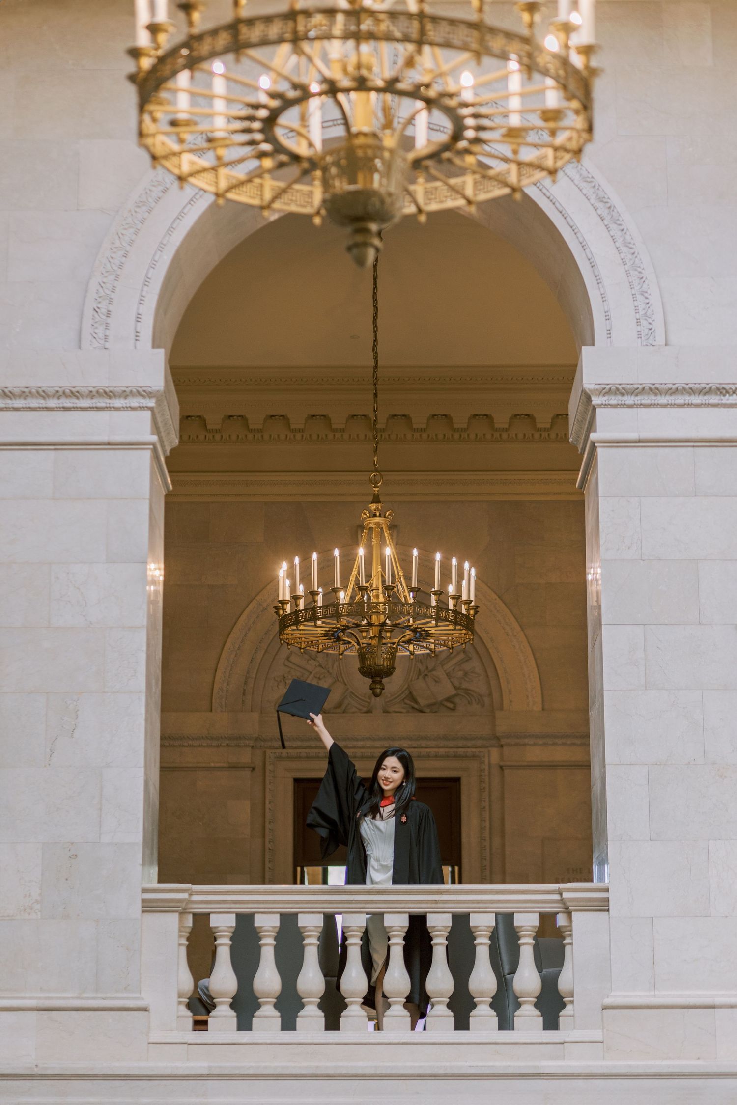 Harvard Graduate in cap and gown raises diploma on ornate balcony beneath grand chandeliers in elegant neoclassical building.