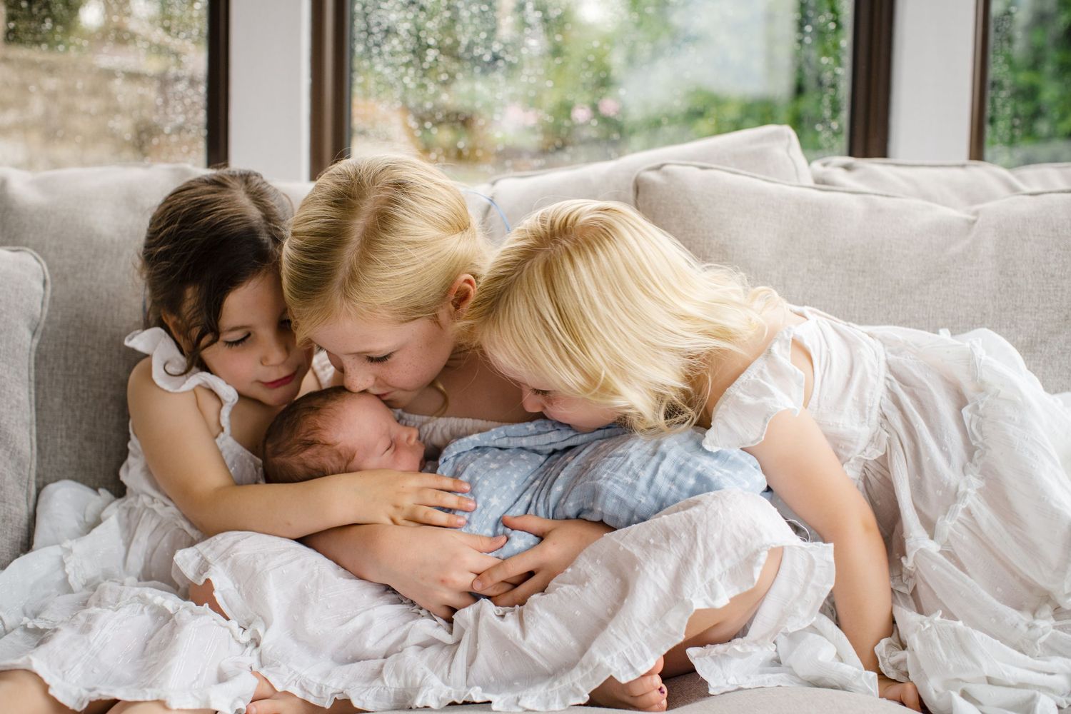 Two older sisters lovingly kiss and cuddle their newborn baby sibling on a cozy couch near a window.