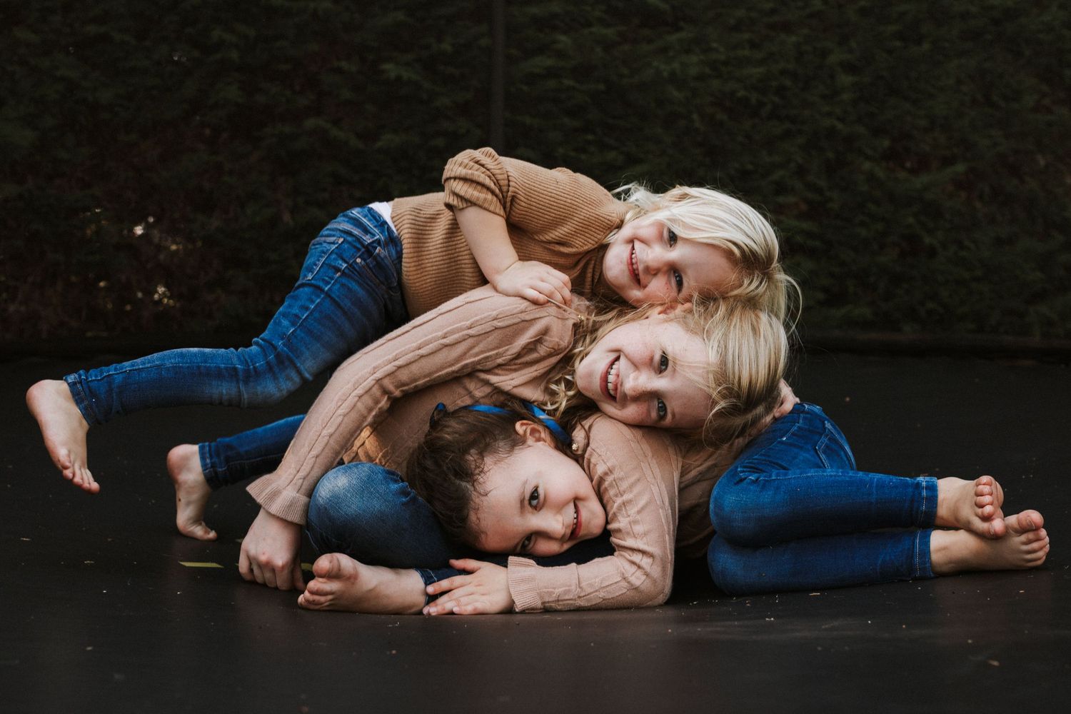 Three children playfully piled on top of each other, smiling and laughing on a dark floor.