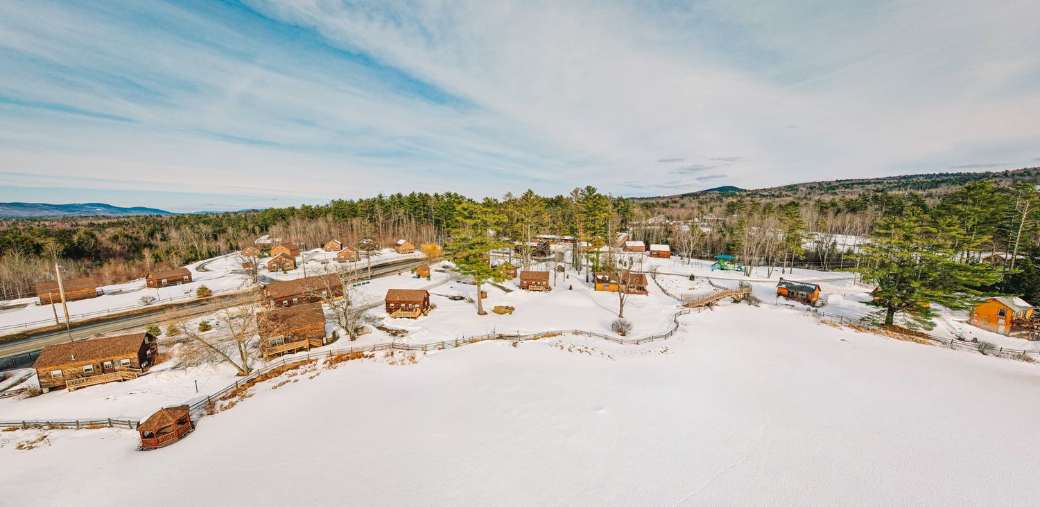Aerial winter panorama of a snowy New England village surrounded by pine forests under blue skies.