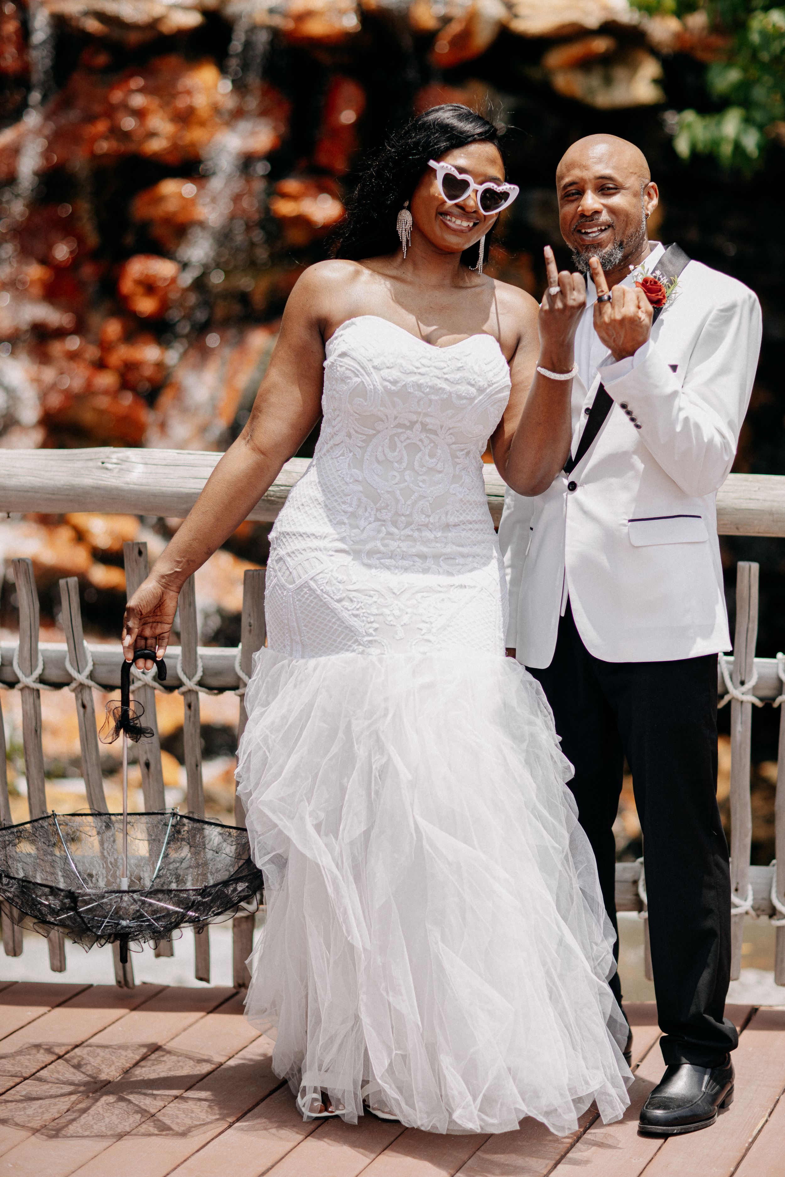 Bride in white mermaid gown with heart sunglasses poses with groom in white tuxedo jacket near fountain.