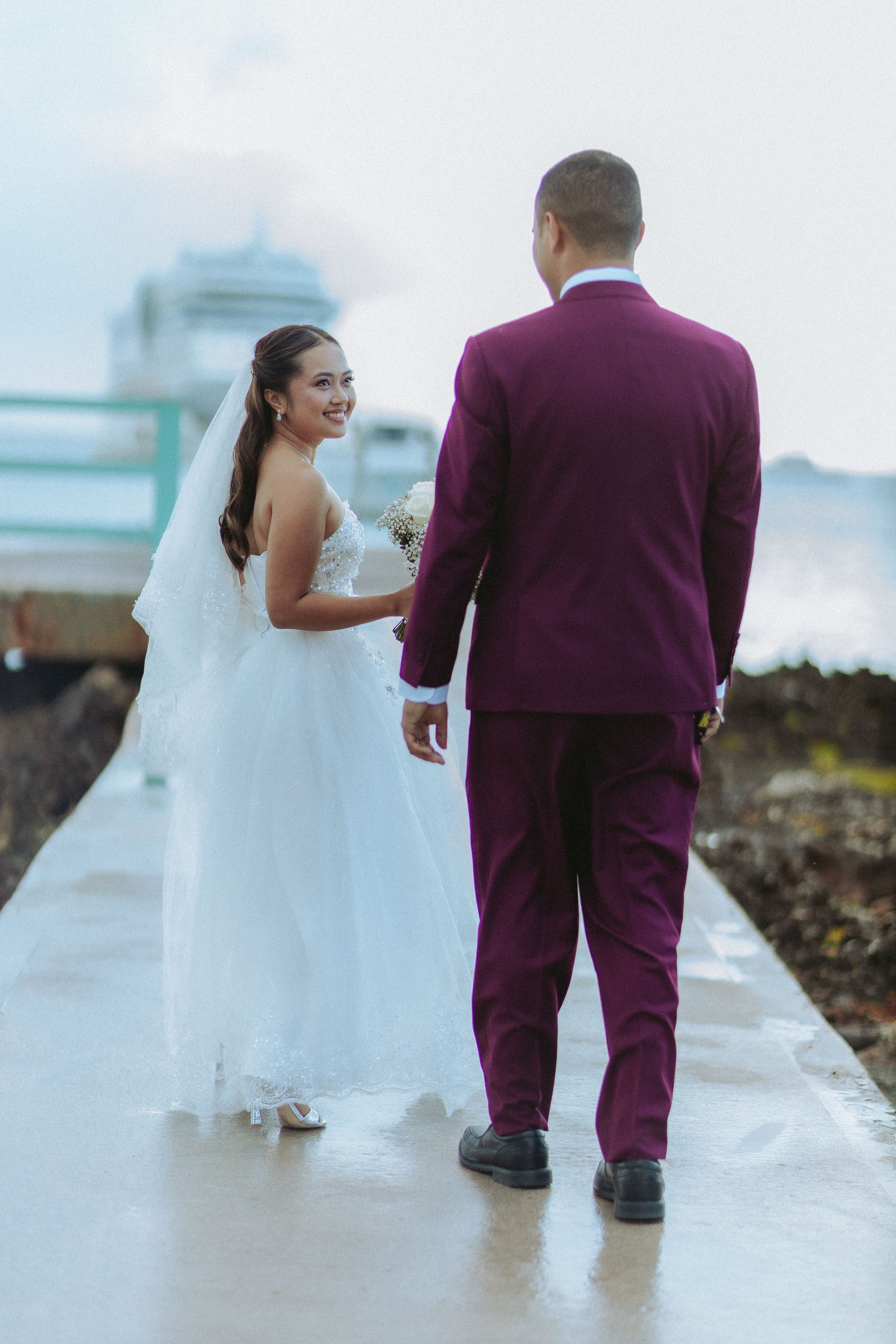Bride in white gown and groom in purple suit walk together on a path, bride smiling back at camera.
