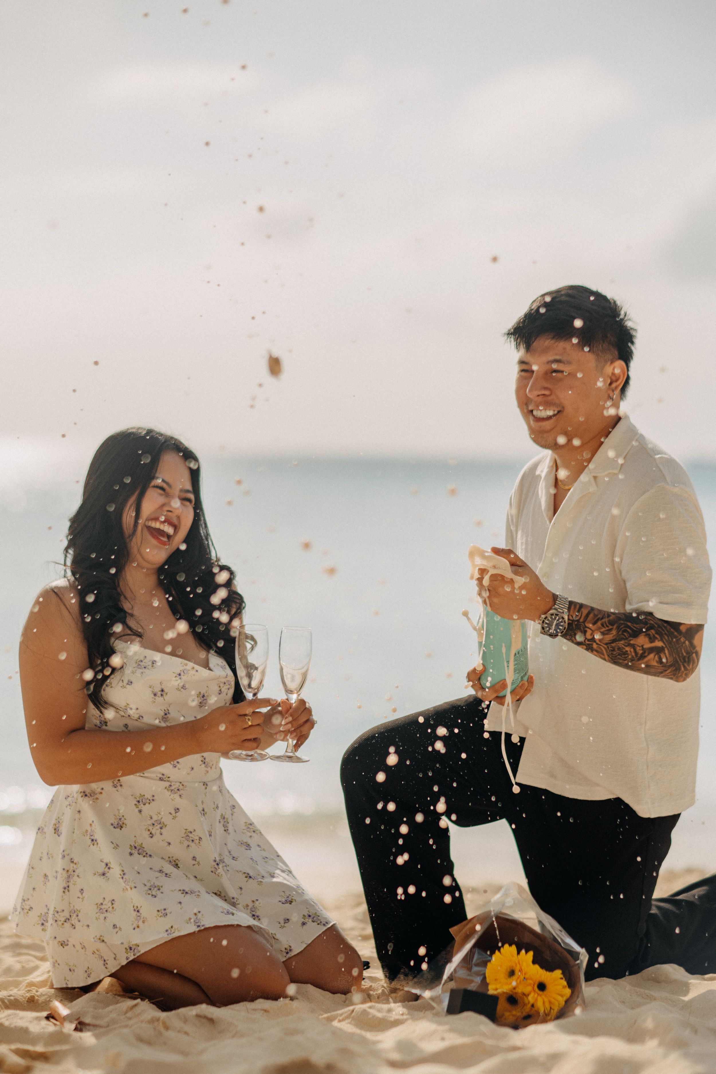 Couple celebrating beach proposal with champagne and confetti, man on one knee with sunflowers on sandy shore.