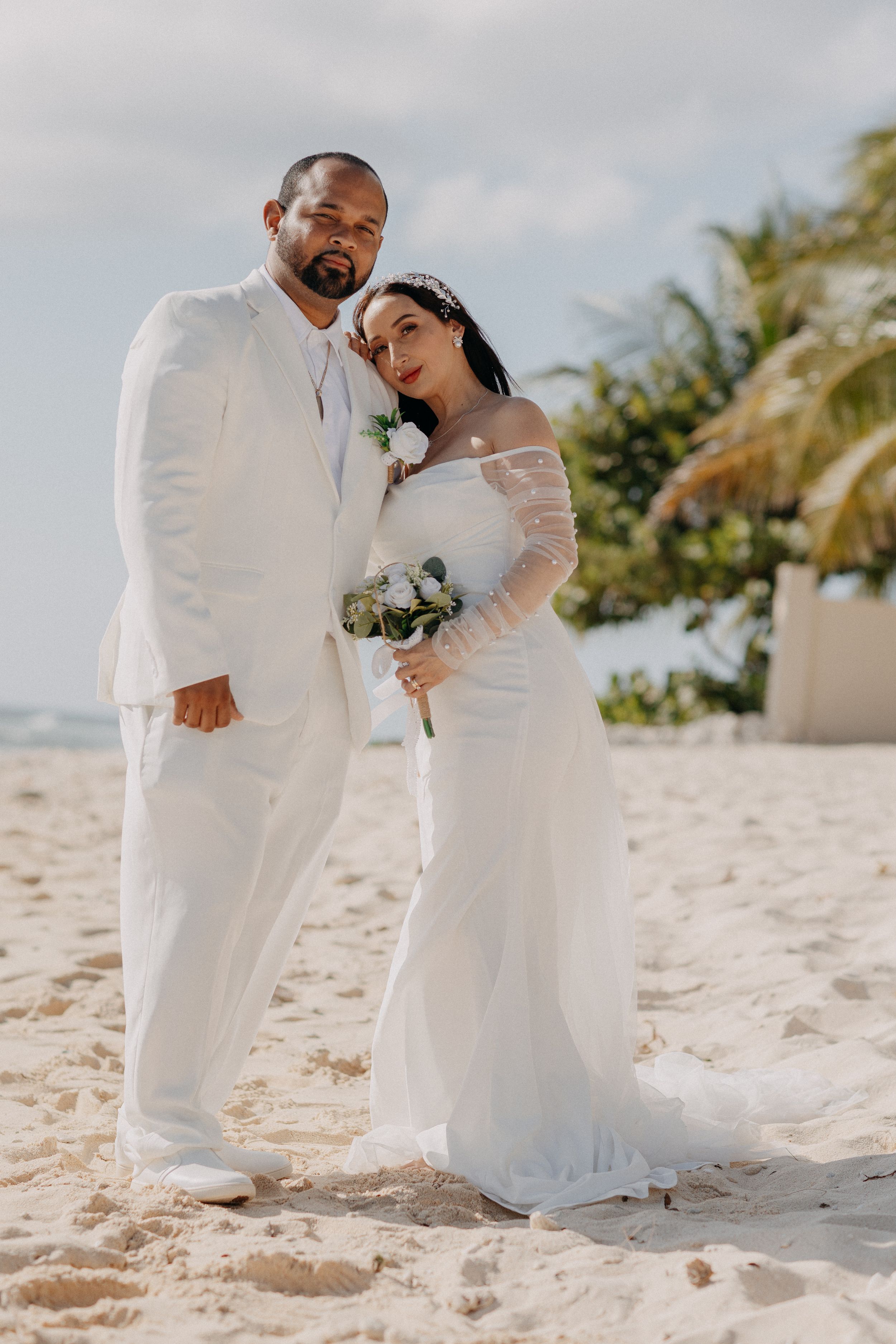 Couple in white wedding attire posing on a sandy beach with palm trees, bride holding a bouquet of white flowers.