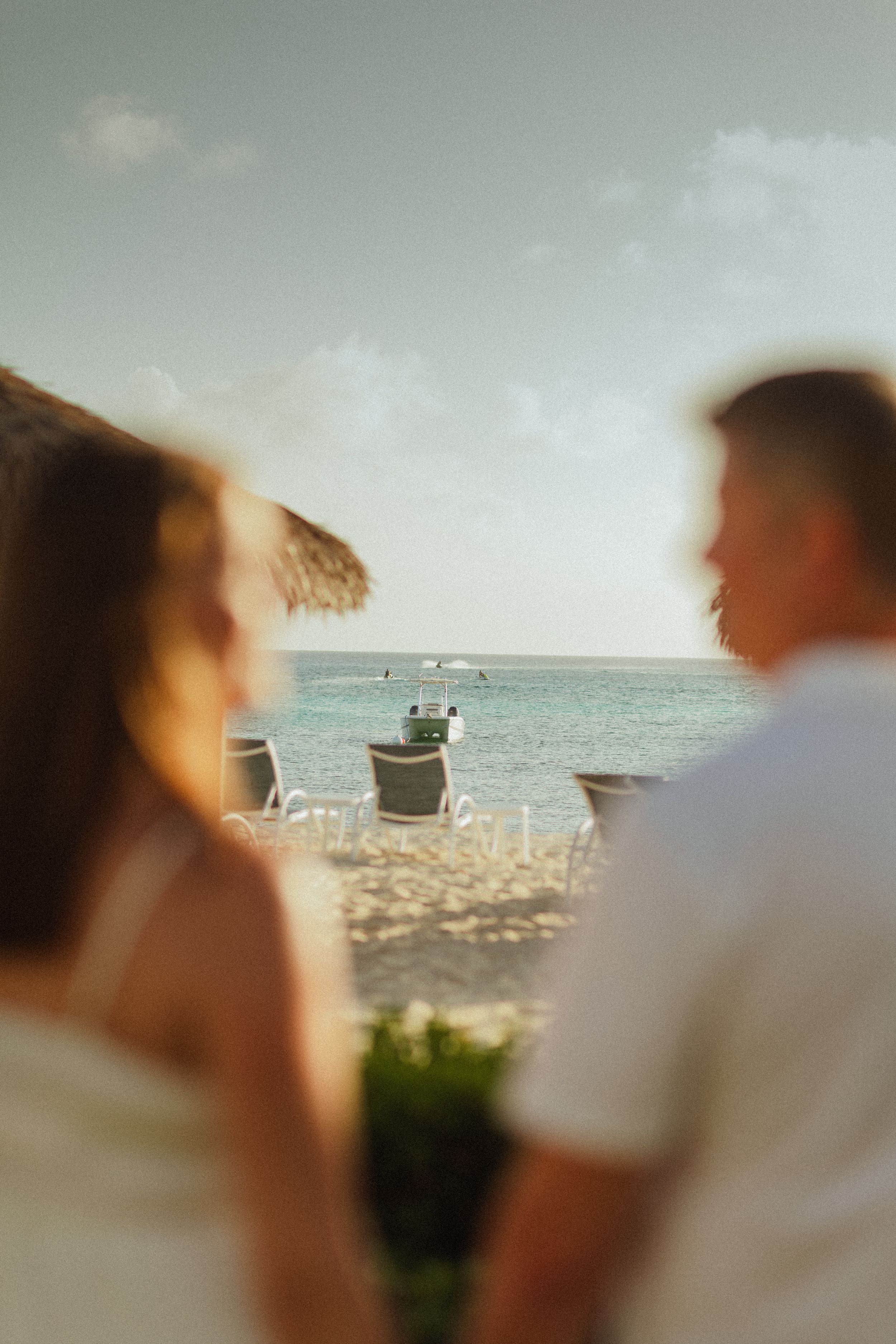 Couple enjoying a romantic beachside view at sunset with turquoise ocean waters and a thatched umbrella in a tropical setting.