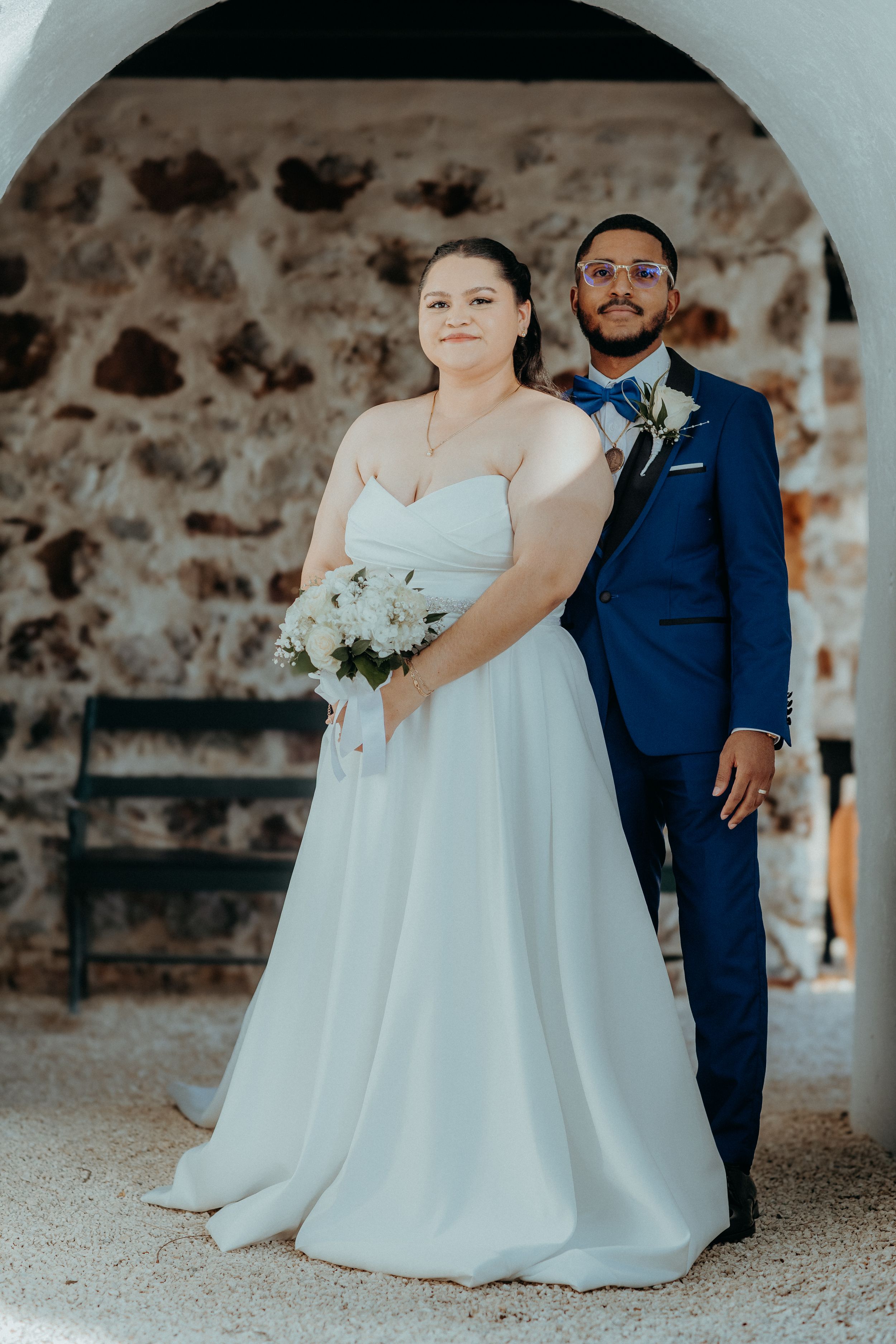 Bride in white gown holding bouquet stands with groom in navy suit under stone arch at wedding venue.