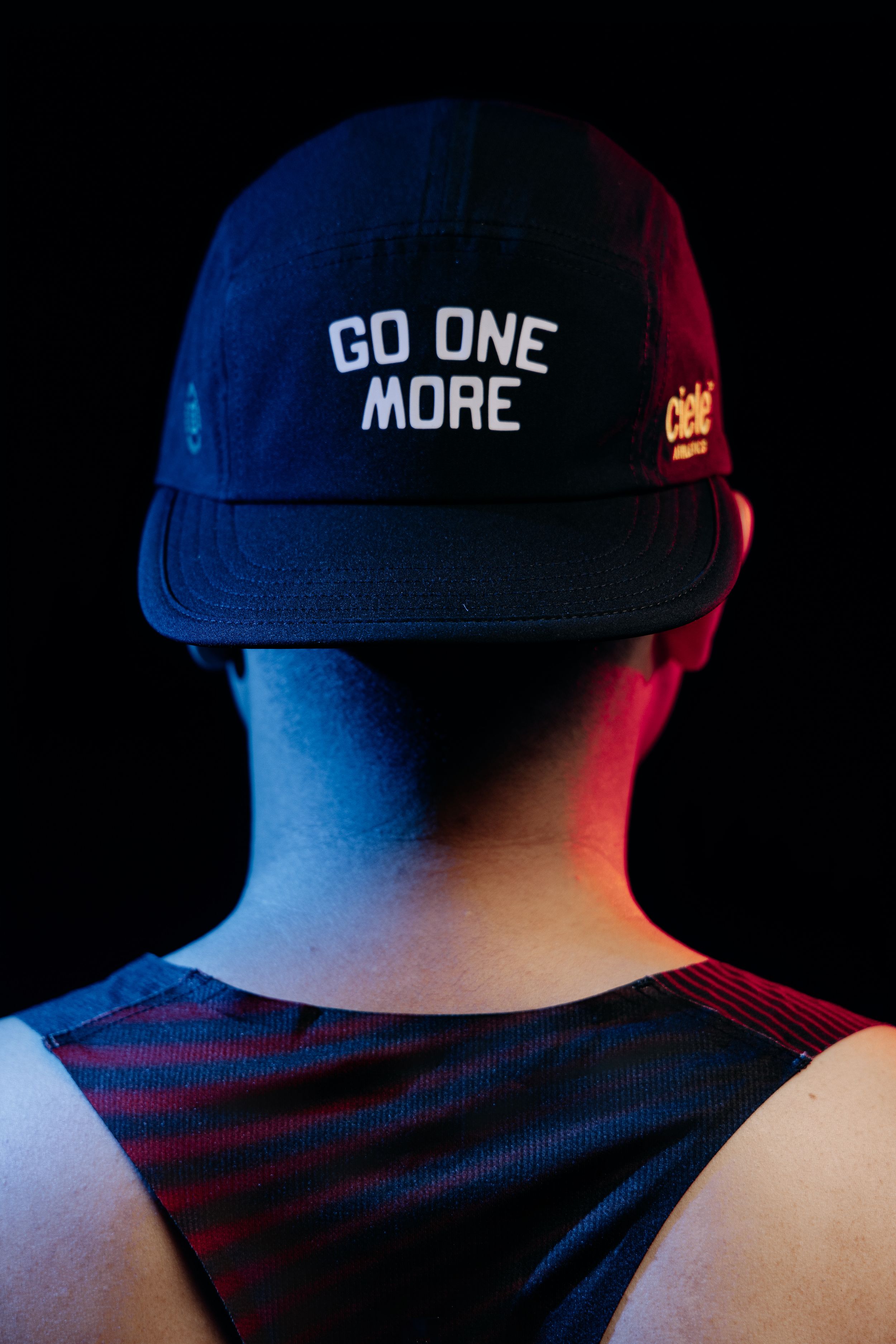 Person wearing a navy Ciele Athletics cap with white 'GO ONE MORE' text, viewed from behind against dark background.