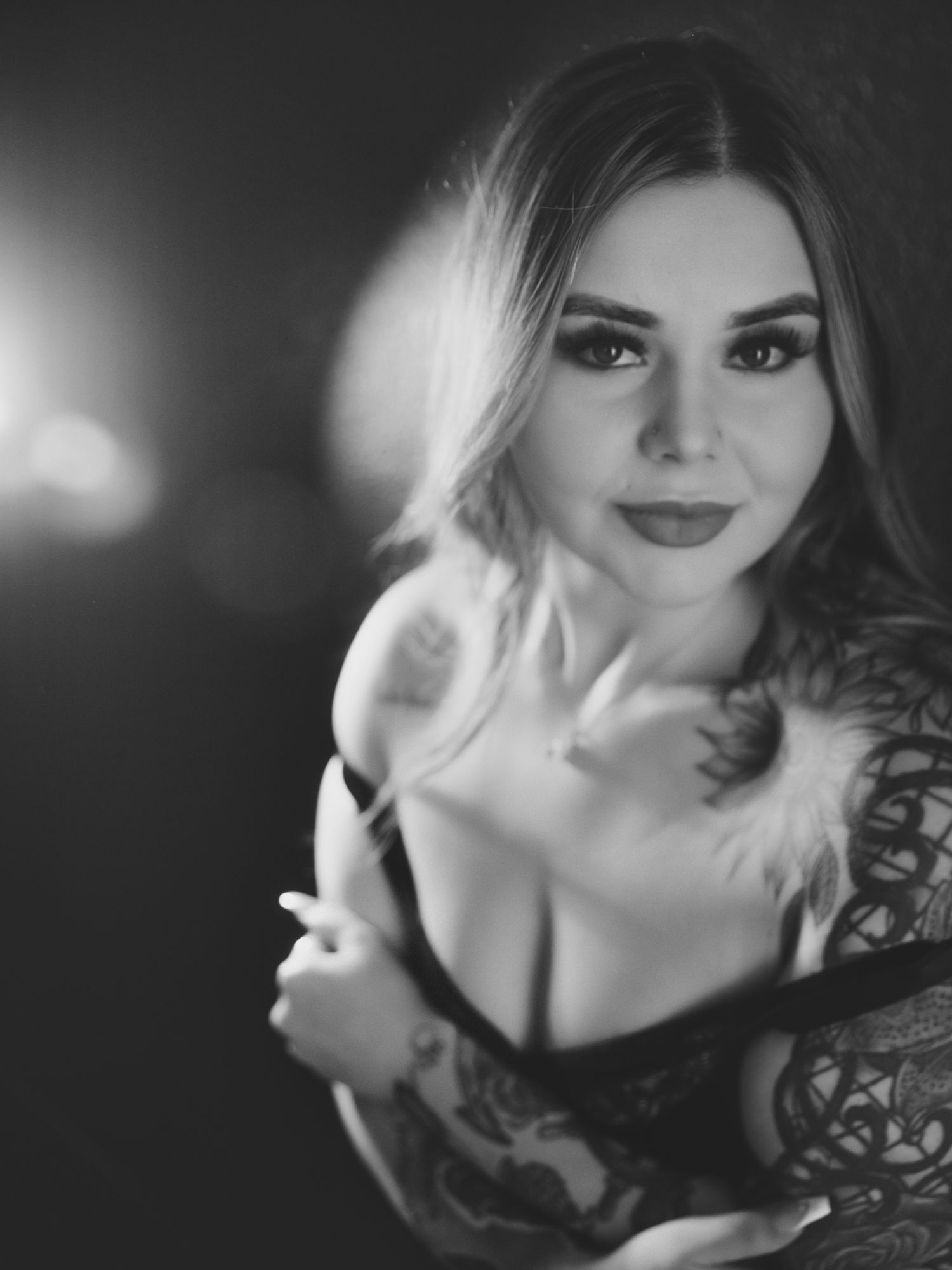 Black and white boudoir portrait of a tattooed woman with bold makeup posing in lingerie with soft bokeh lighting.