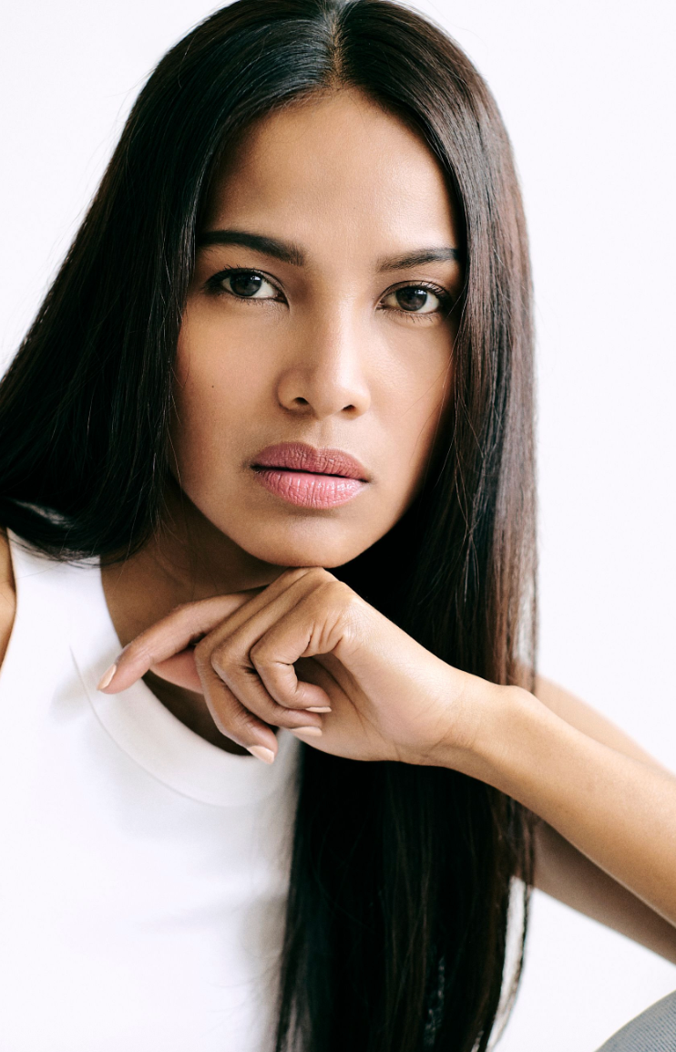 Young woman with long straight black hair poses confidently in white top against light background.