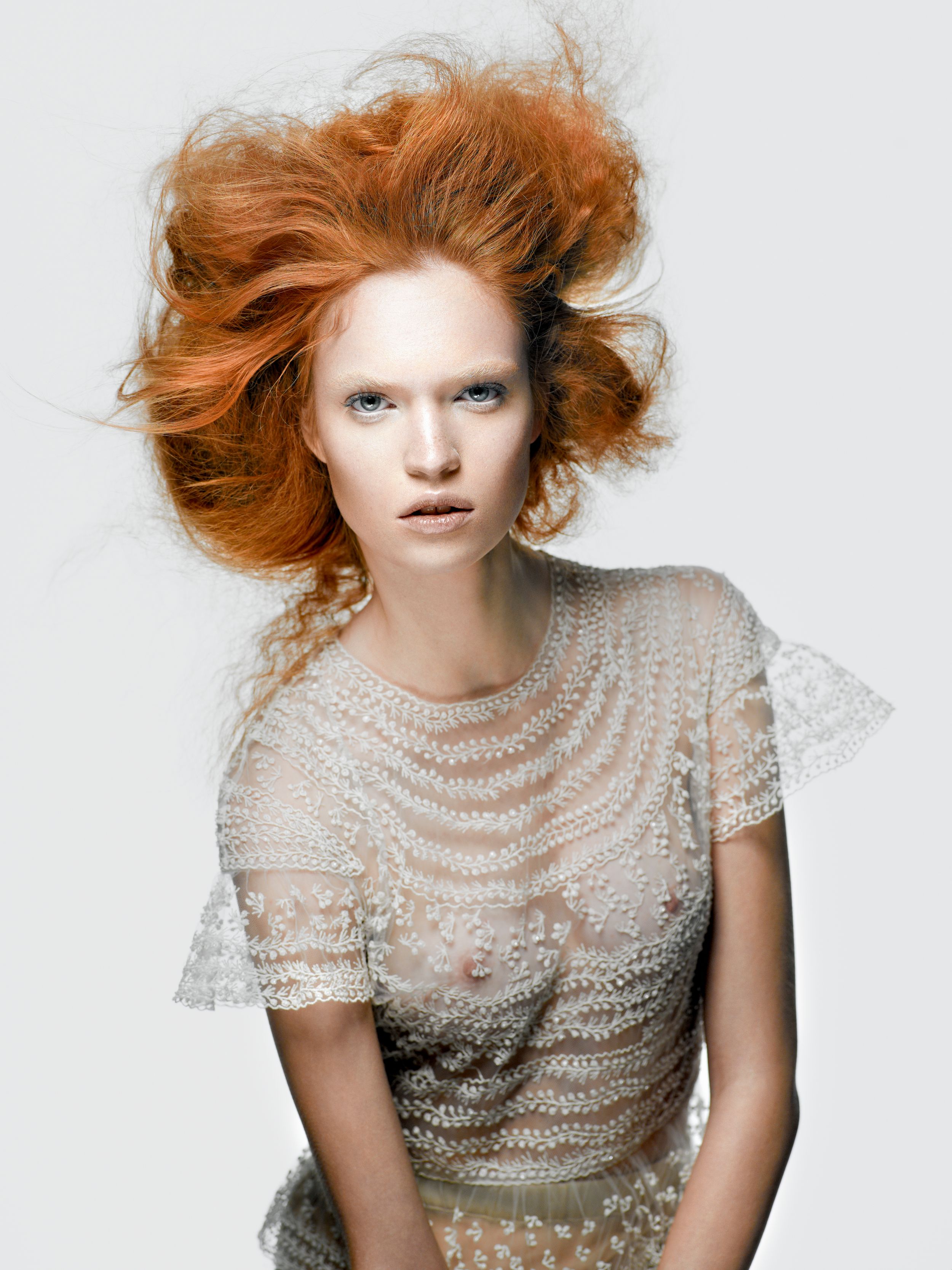 A model with voluminous red hair wearing an elegant white lace embellished top against a light background.