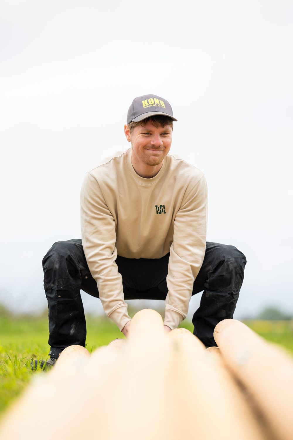 Man in beige sweatshirt and cap smiling while handling wooden logs outdoors on green grass.