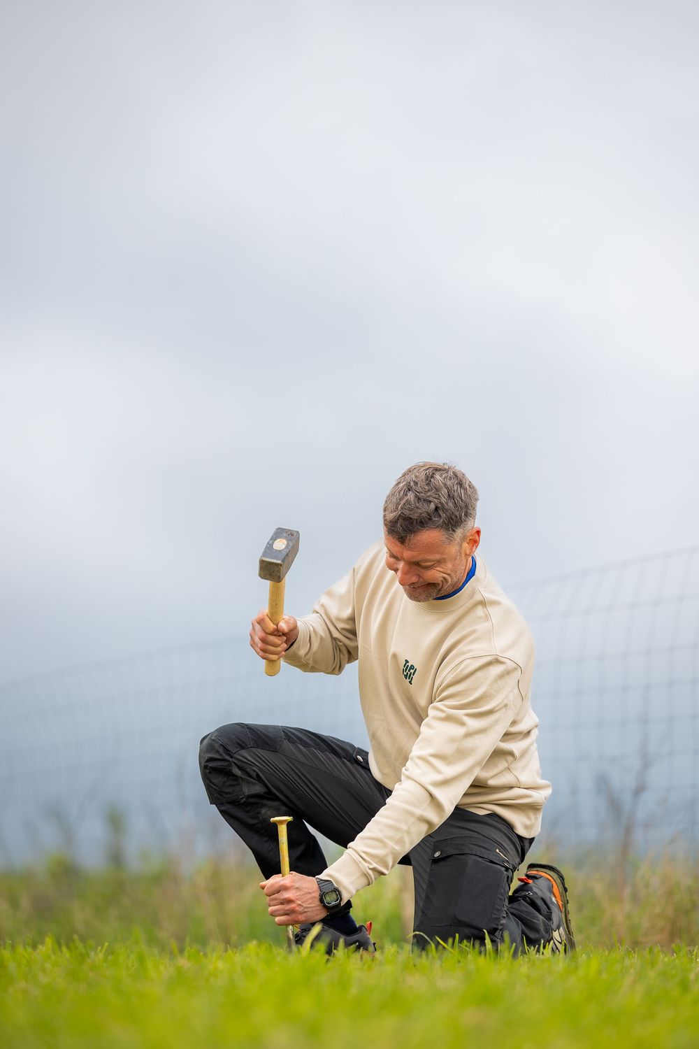 Man kneeling on grass driving a stake into the ground with a wooden mallet on a cloudy day.