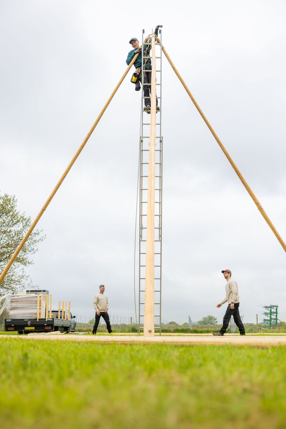 Workers erecting a tall metal ladder structure with wooden support poles on a green field under cloudy skies.