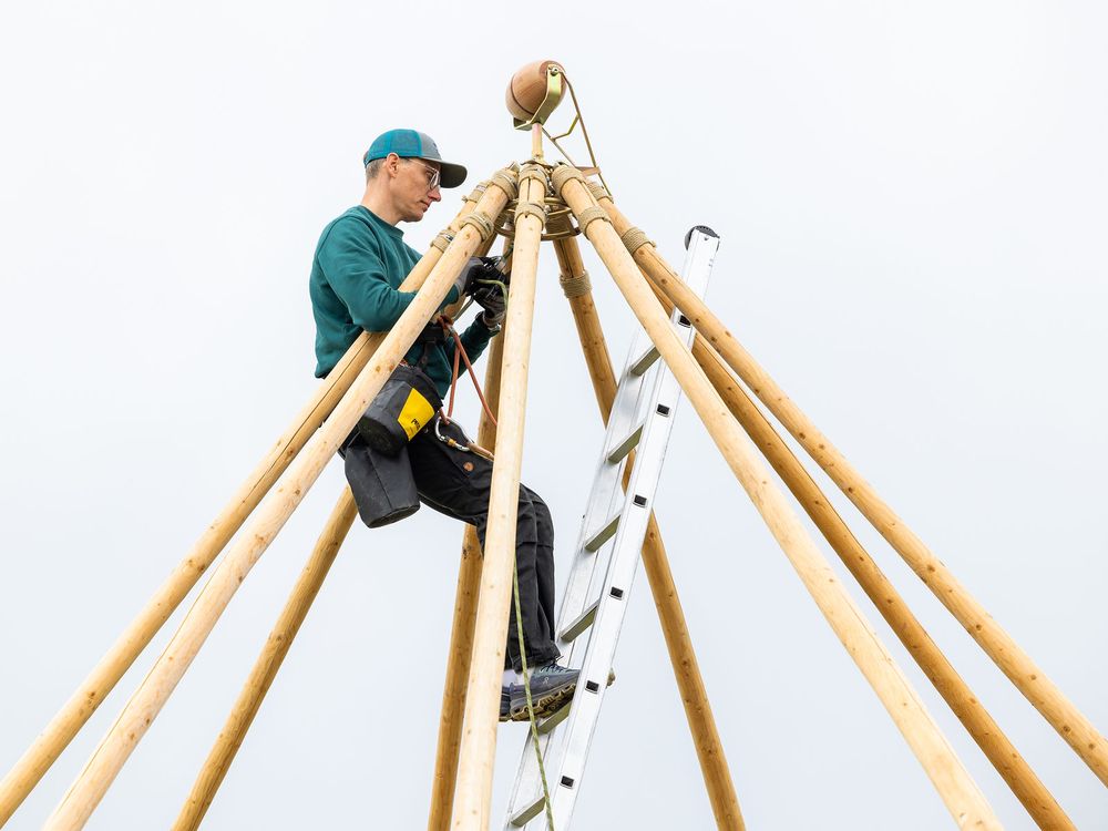Worker climbing a ladder on a wooden pole structure against a cloudy sky.
