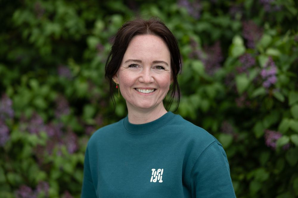 Smiling woman in teal sweatshirt posing outdoors in front of lush green and purple flowering bushes.