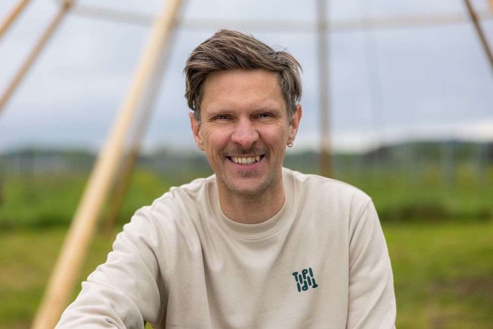 Smiling man in beige sweatshirt outdoors near wooden structure with green landscape background.