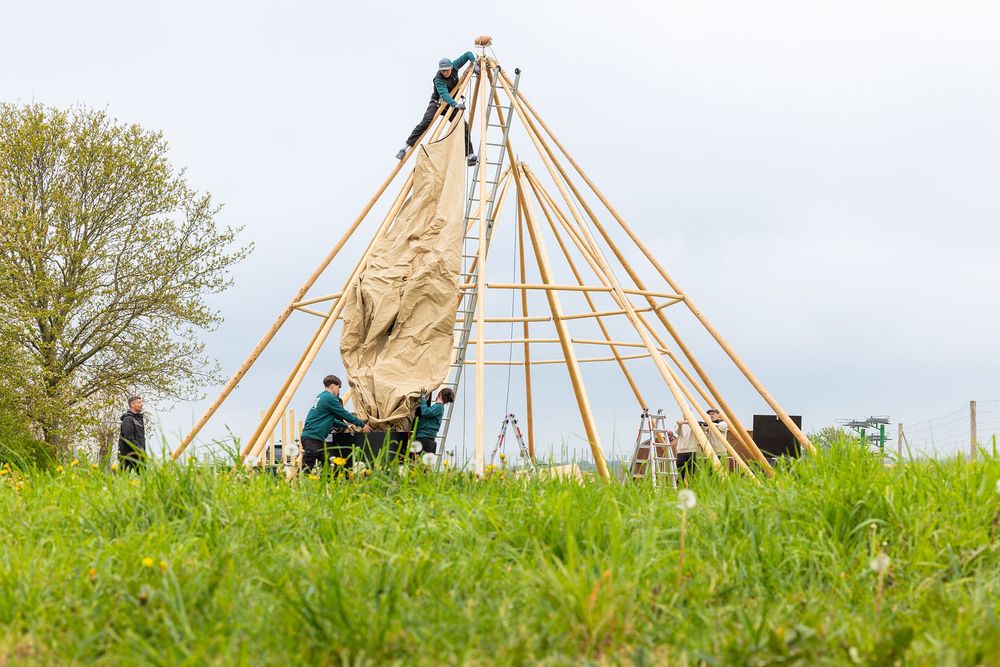 Group of people constructing a large wooden teepee frame with canvas covering in an open green field.