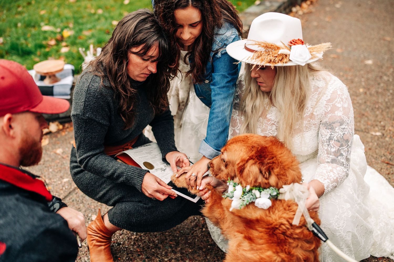 Bride in white lace dress and hat signs document with golden retriever wearing flower collar at outdoor wedding ceremony.