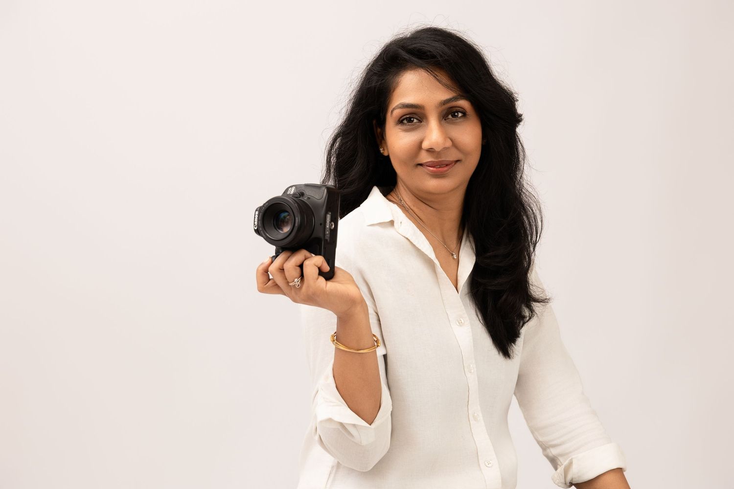 Woman in white shirt holding a DSLR camera, smiling confidently against a light background.