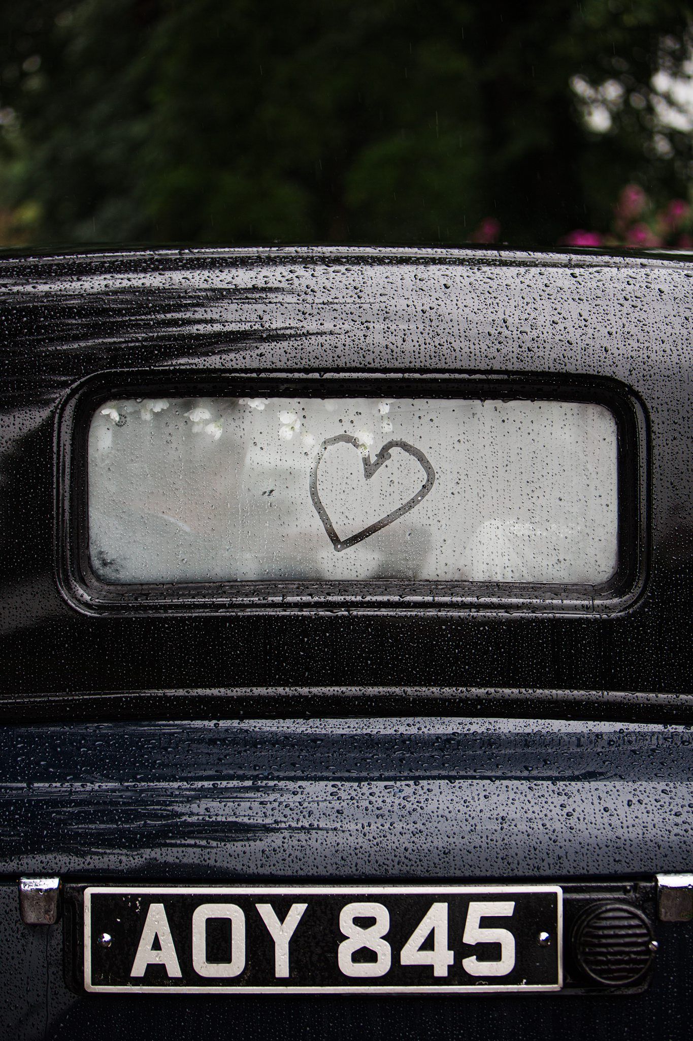 Heart drawn on foggy rear window of vintage black car with license plate AOY 845, covered in rain droplets.