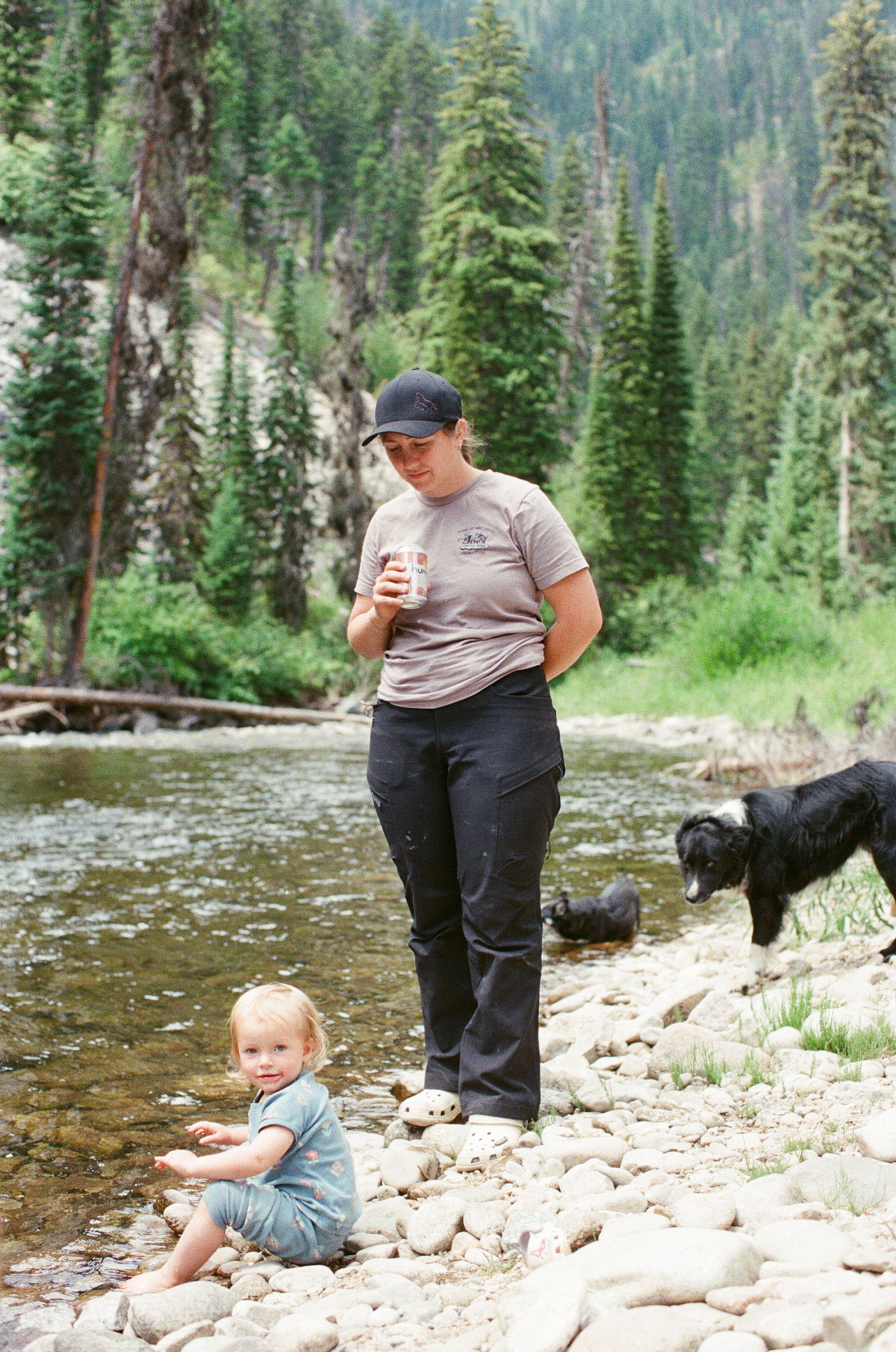 Woman and toddler by rocky mountain stream with black dog, surrounded by pine forest.