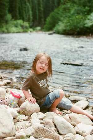 Young child sitting on rocky riverbank wearing Vans t-shirt and jeans, smiling outdoors near a forest stream.